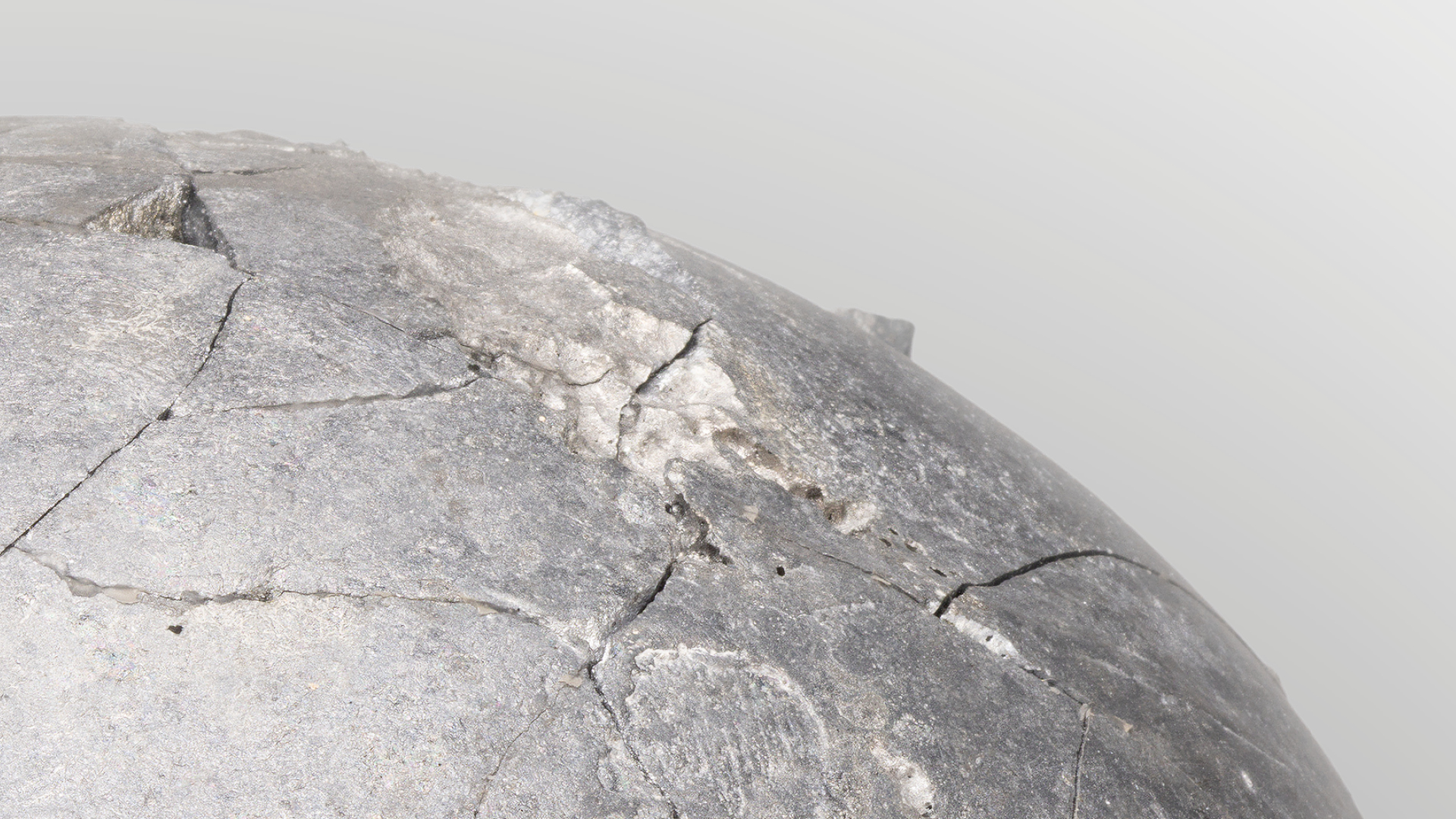 Close-up of a piece of artwork which is a dry crusty surface of a clay sphere, painted in a metallic sliver colour, in front of a grey-coloured background.